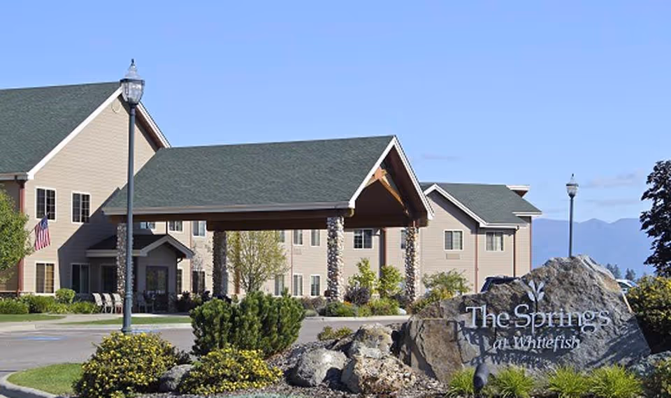 Front entrance of The Springs at Whitefish senior living building with a covered porte-cochere, landscaped rock sign, and lampposts.