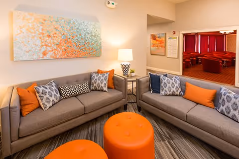 Cozy seating area with two gray sofas, orange ottomans and patterned pillows in a lounge with wall art and a lamp.