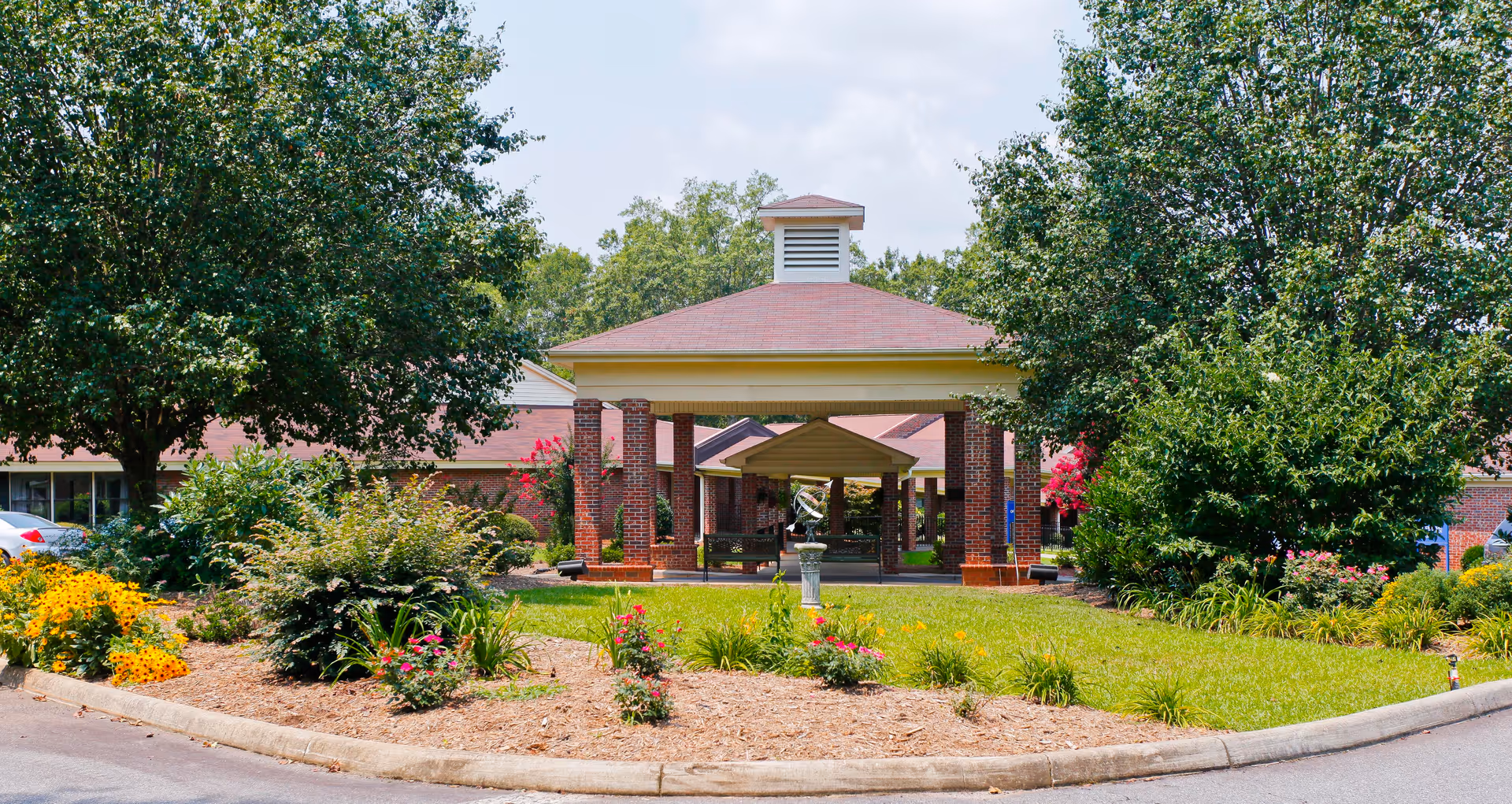 Front entrance of The Renaissance Senior Living facility featuring a covered driveway with brick pillars, surrounded by well-maintained landscaping including green bushes, colorful flowers, and trees under a partly cloudy sky.