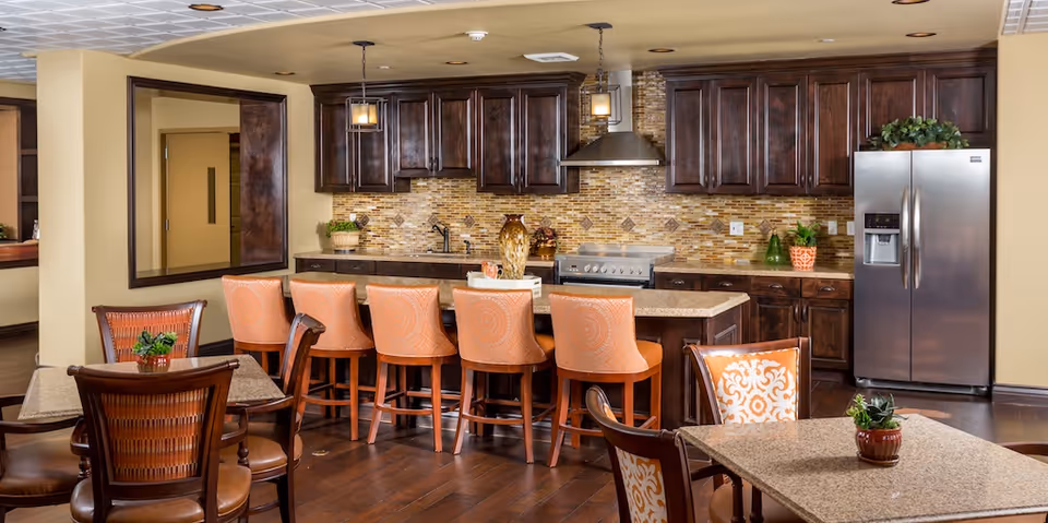 A spacious kitchen and dining area with dark wooden cabinets, a tiled backsplash, and a stainless steel refrigerator. There is a large island with a granite countertop and six orange cushioned bar stools. In the foreground, there are dining tables with chairs, some with patterned cushions and small potted plants on the tables.
