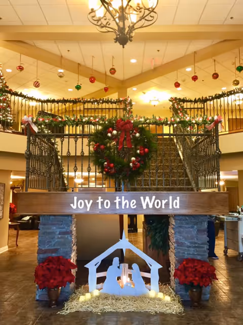 Festively decorated interior lobby with a 'Joy to the World' sign, nativity scene, wreaths, garlands, and a central staircase.