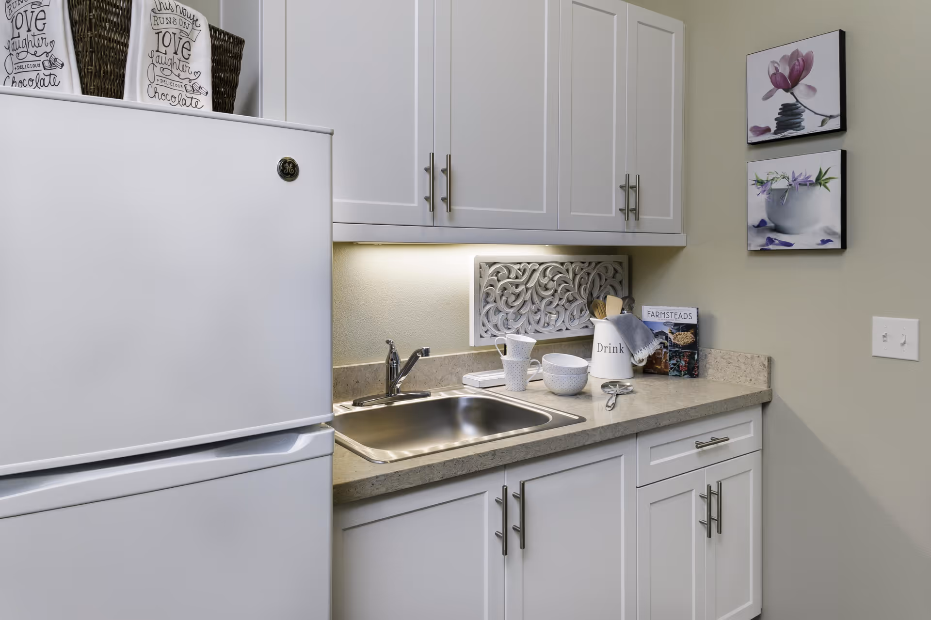 Compact kitchenette with white cabinets, a stainless steel sink, countertop accessories, and a white refrigerator.