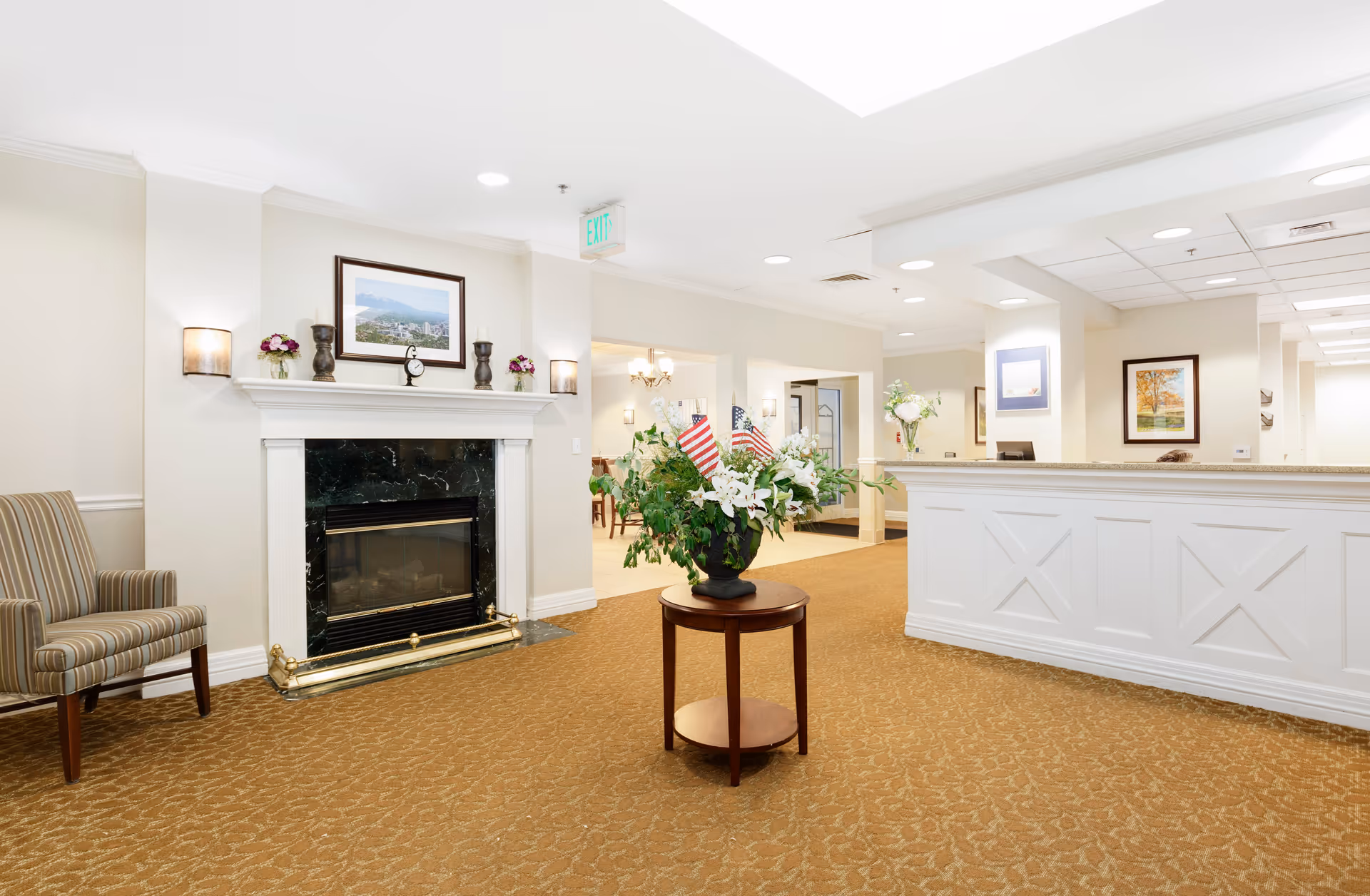A bright and welcoming assisted living facility lobby with a white reception desk on the right, a small round table with a floral arrangement and American flags in the center, a striped armchair on the left, and a fireplace with a framed picture above it on the left wall. The space has beige walls, carpeted floors, and recessed ceiling lights.
