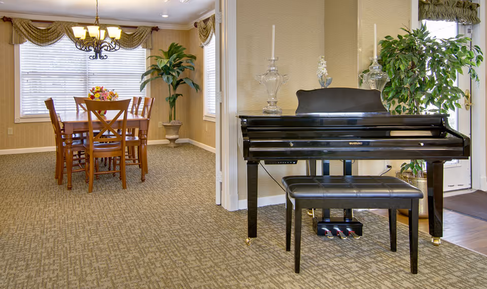 Interior view of a senior living facility showing a dining area with a wooden table and six chairs, a chandelier above the table, a large window with blinds and valances, a potted plant in the corner, and a black Suzuki piano with a matching bench near the entrance door.