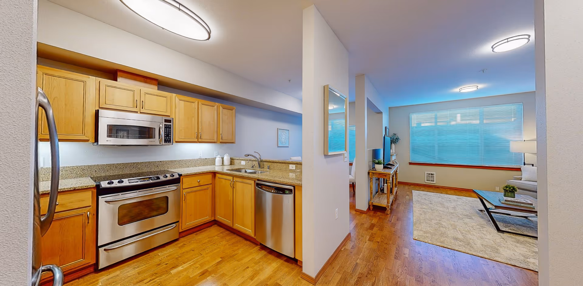 Interior view of a senior living facility apartment showing a kitchen with wooden cabinets, stainless steel appliances including a microwave, oven, dishwasher, and refrigerator. Adjacent to the kitchen is a living room area with a large window, a rug, a coffee table, a TV on a wooden stand, and a sofa.