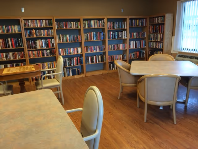 A cozy room with wooden flooring featuring multiple bookshelves filled with books along the walls. There are several tables with beige cushioned chairs arranged around them, and a window with curtains letting in natural light.