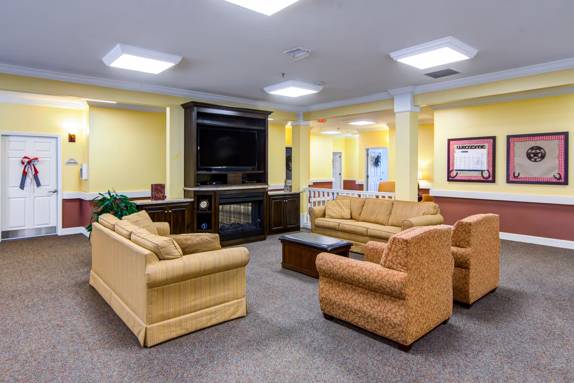 A cozy living room area in a senior living facility with two beige sofas, two patterned armchairs, a dark wooden coffee table, and a large dark wooden entertainment center with a TV and electric fireplace. The walls are painted yellow with white trim, and there are framed decorations on the wall. The carpet is a neutral tone, and the ceiling has recessed lighting.