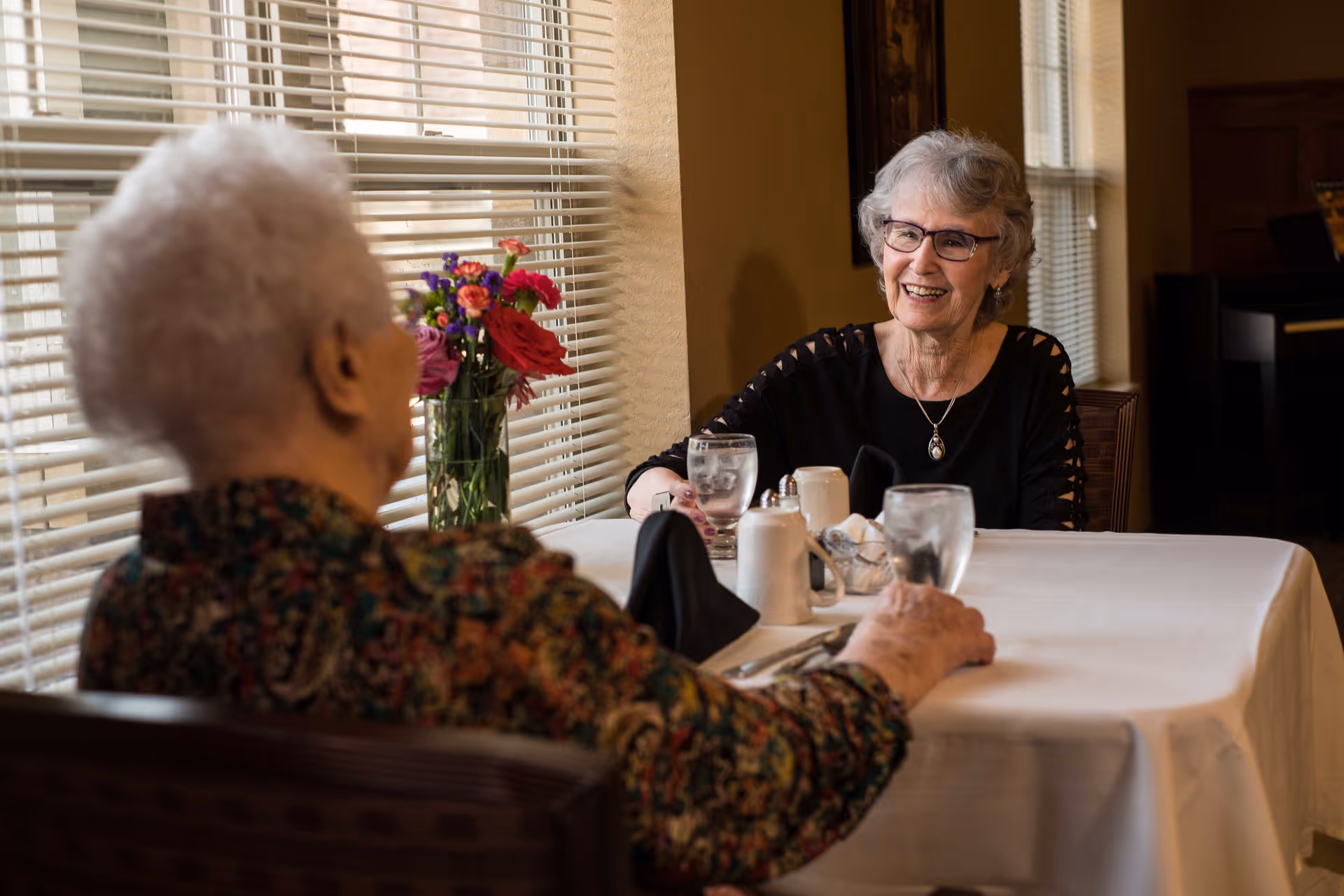 Two elderly women sitting at a dining table covered with a white tablecloth, engaged in conversation. One woman is smiling and wearing glasses, while the other has her back to the camera. There is a vase with colorful flowers on the windowsill behind them and sunlight coming through the blinds.