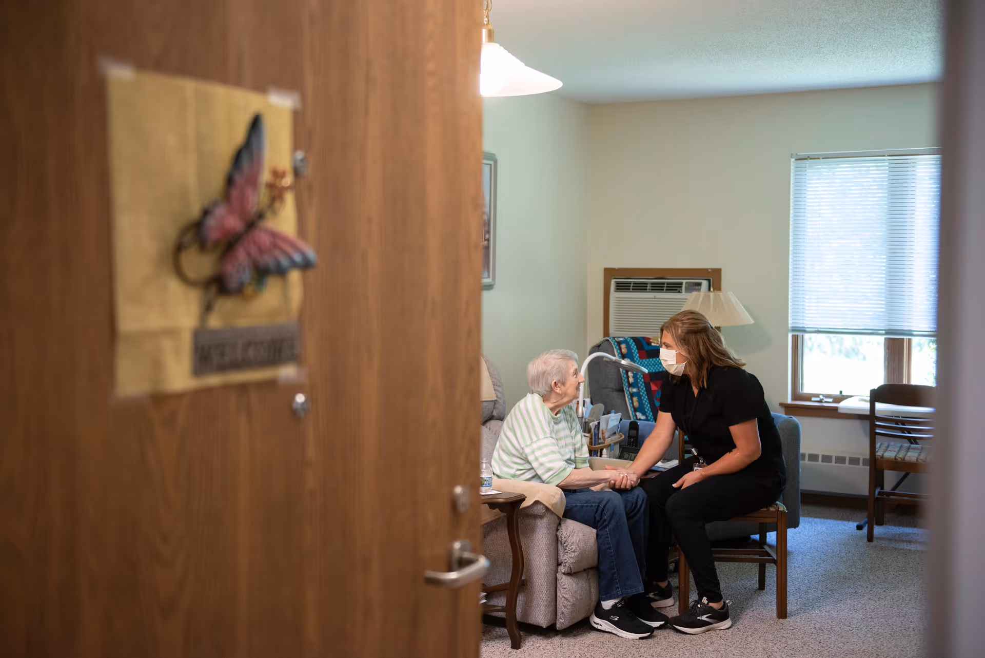 View through a partially open door into a cozy room where an elderly woman sits on a recliner chair holding hands and talking with a caregiver wearing a face mask who is seated on a wooden chair. The room has a window with blinds, an air conditioning unit, a lamp, and a small table beside the recliner.
