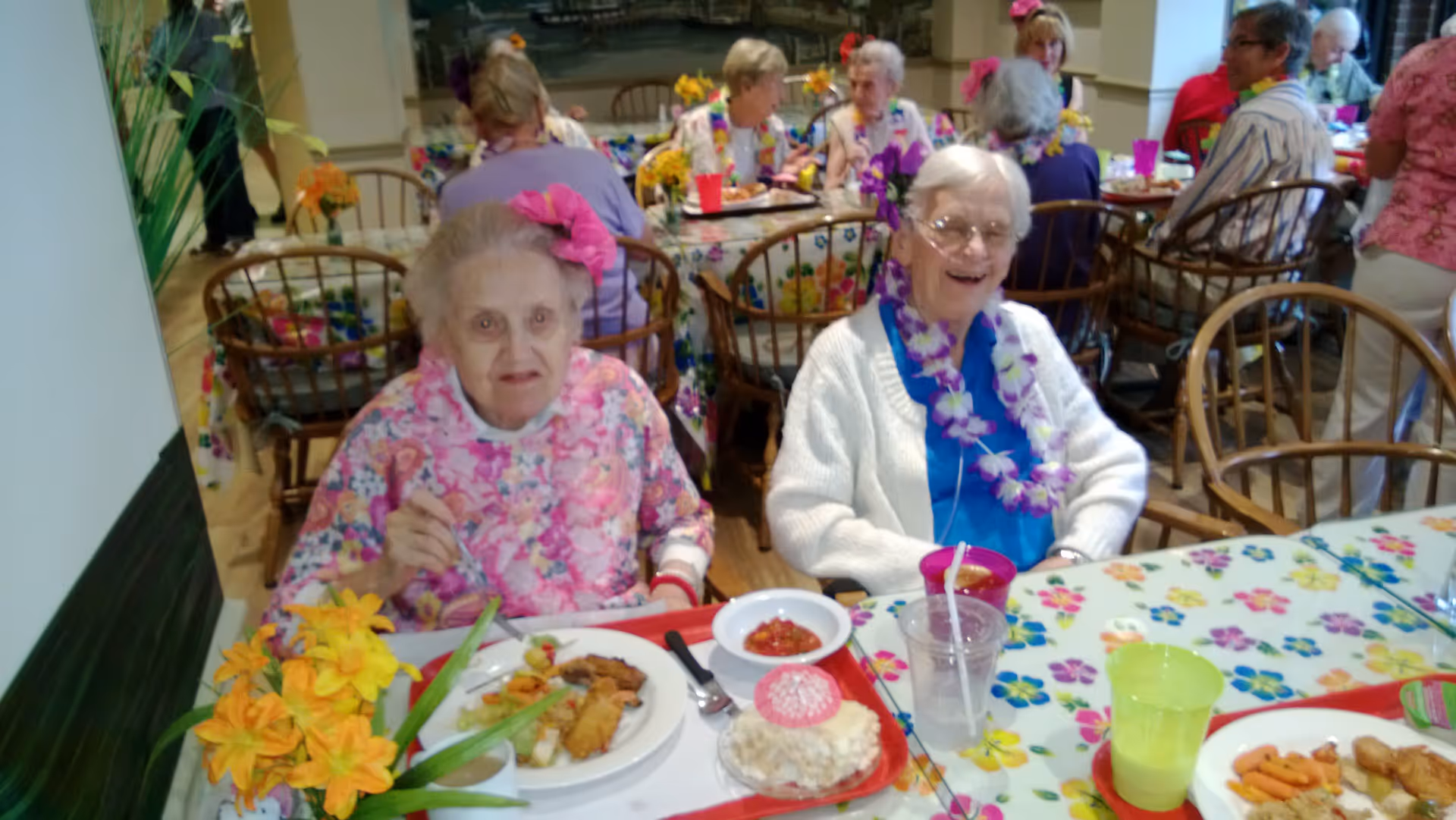 Two elderly women sitting at a table in a dining area, enjoying a meal. One woman is wearing a pink floral top with a pink flower in her hair, and the other is wearing a white sweater with a blue shirt and a purple lei around her neck. Other elderly people are seated at tables in the background, with colorful floral tablecloths and decorations.