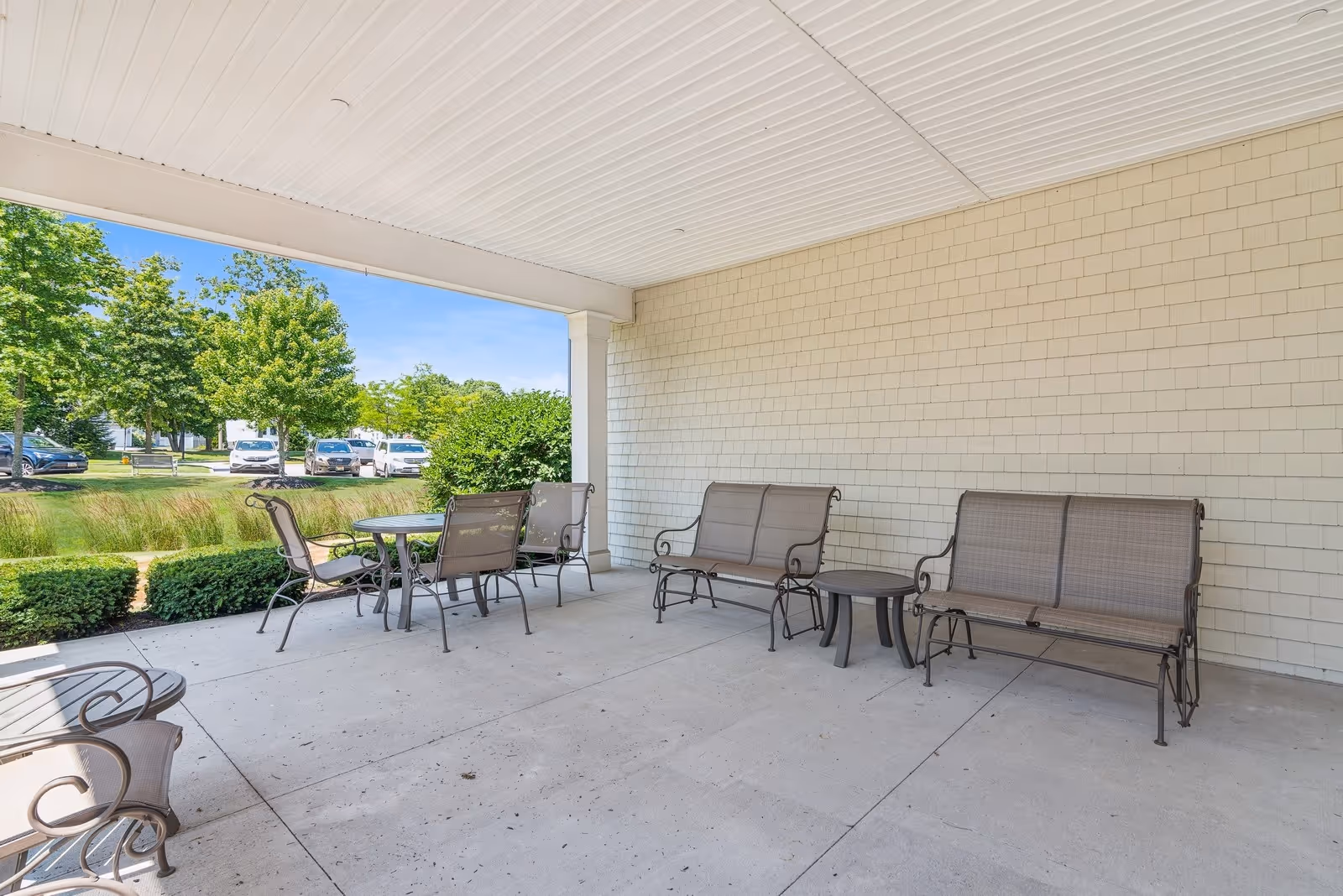 Covered outdoor patio with metal tables, chairs and benches overlooking landscaped grounds and parked cars.