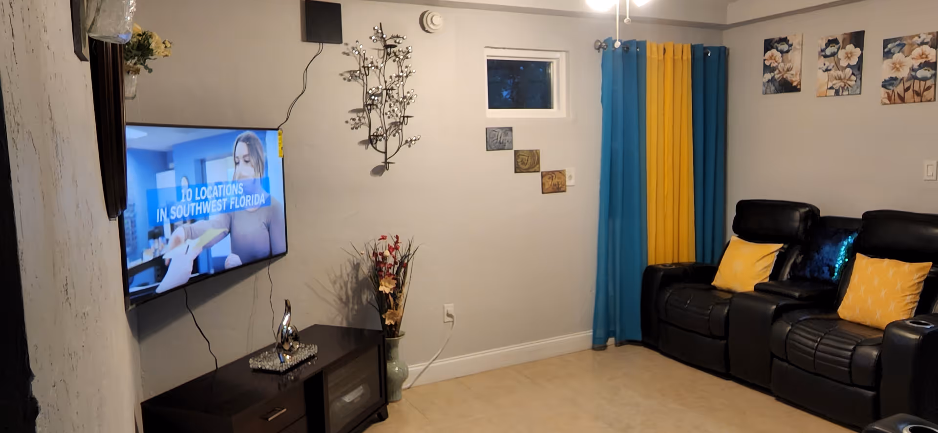A cozy living room with a wall-mounted flat screen TV displaying a program about locations in Southwest Florida. Below the TV is a dark wooden cabinet with decorative items. On the right side, there are two black leather recliners with yellow and blue pillows. The room has light-colored walls with decorative wall art and a window with blue and yellow curtains.
