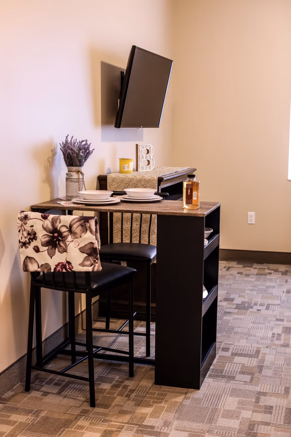 A small dining area with a high table set for two, featuring two black chairs with floral-patterned cushions. On the table are two white bowls and plates, and a bottle of liquid. Behind the table is a cabinet with a decorative runner, a yellow candle, and a decorative letter B. A flat-screen TV is mounted on the wall above the cabinet. The room has beige walls and patterned carpet flooring.
