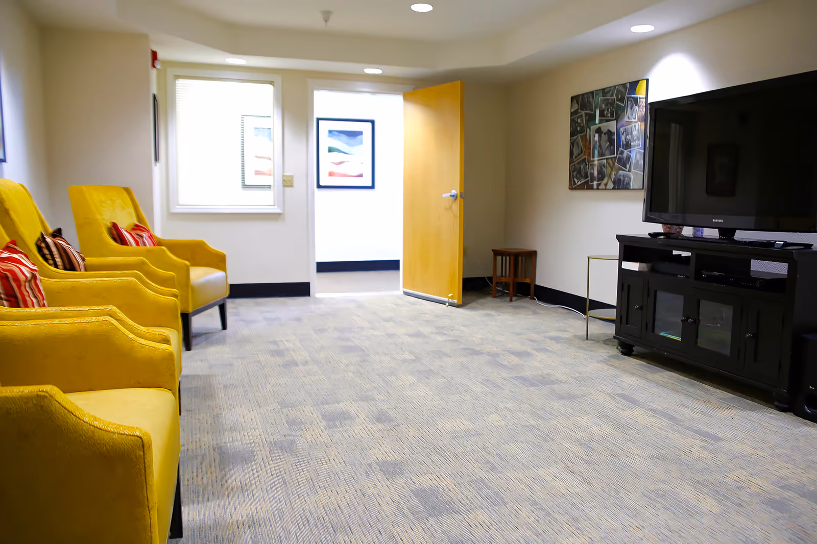 A senior living facility common room with three yellow armchairs with striped pillows on the left side, a large flat-screen TV on a black TV stand on the right, a small wooden stool and a round side table next to the TV stand, beige walls with framed pictures, a window with blinds, and an open wooden door leading to a hallway with a framed picture on the wall.