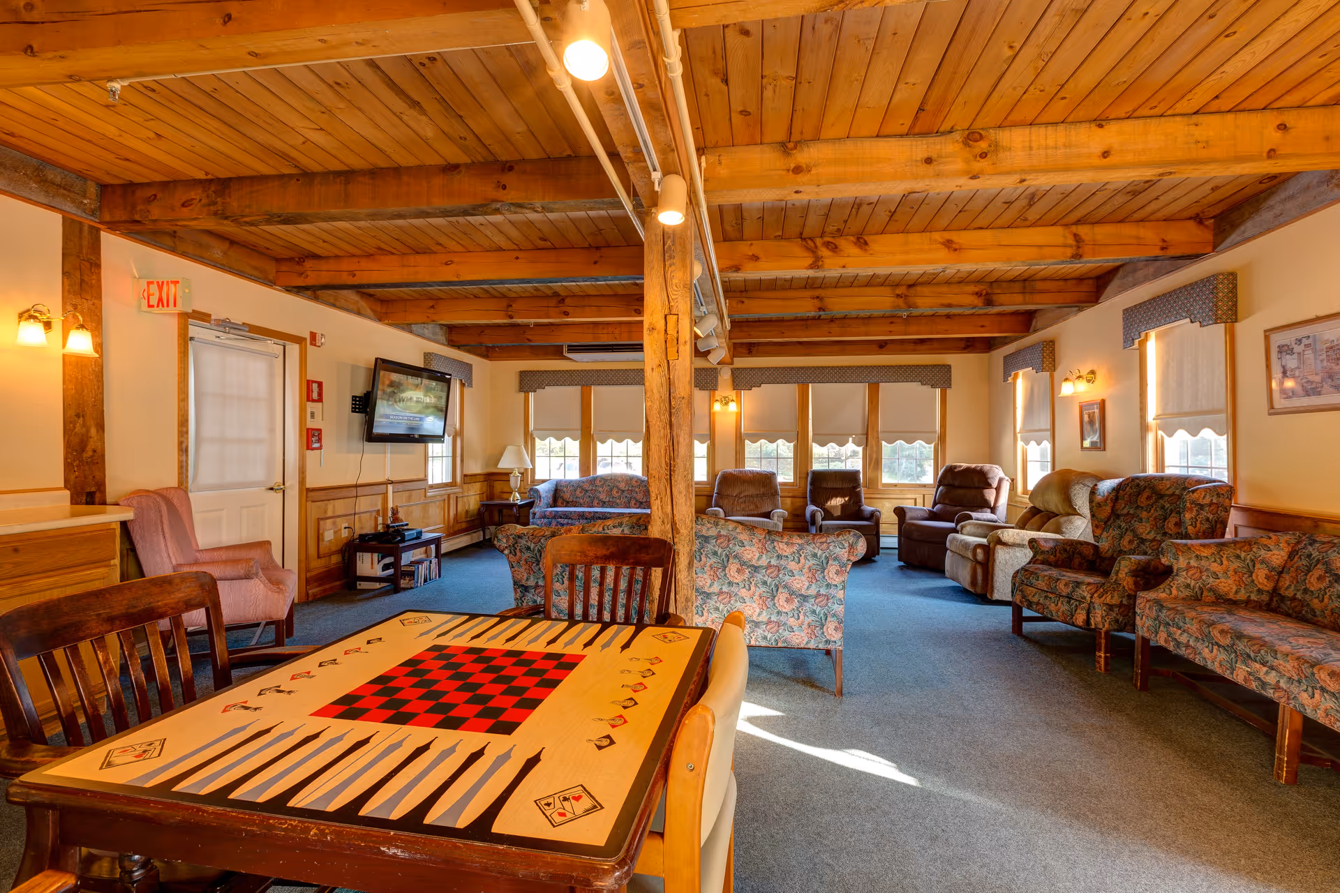 A cozy common living area with wooden ceiling beams and multiple upholstered armchairs and sofas arranged around the room. A table with a built-in checkerboard and backgammon game is in the foreground. The room is well-lit with natural light coming through several windows with valances and shades. A flat-screen TV is mounted on the wall near an exit door.