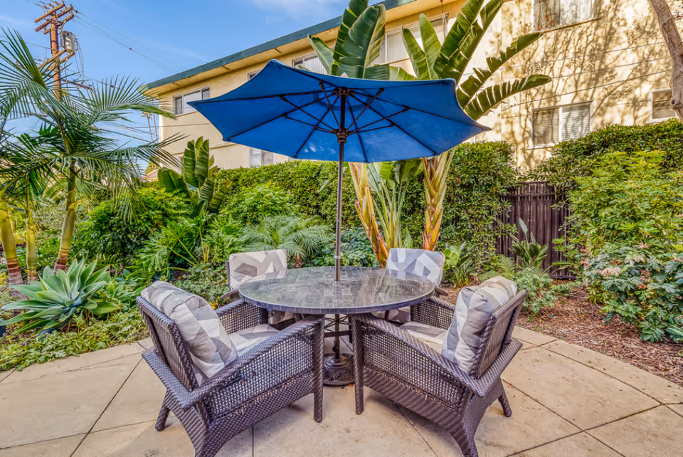Outdoor patio area with a round glass table surrounded by four wicker chairs with cushions, shaded by a large blue umbrella. The patio is surrounded by lush green plants and trees, with a building visible in the background under a clear blue sky.