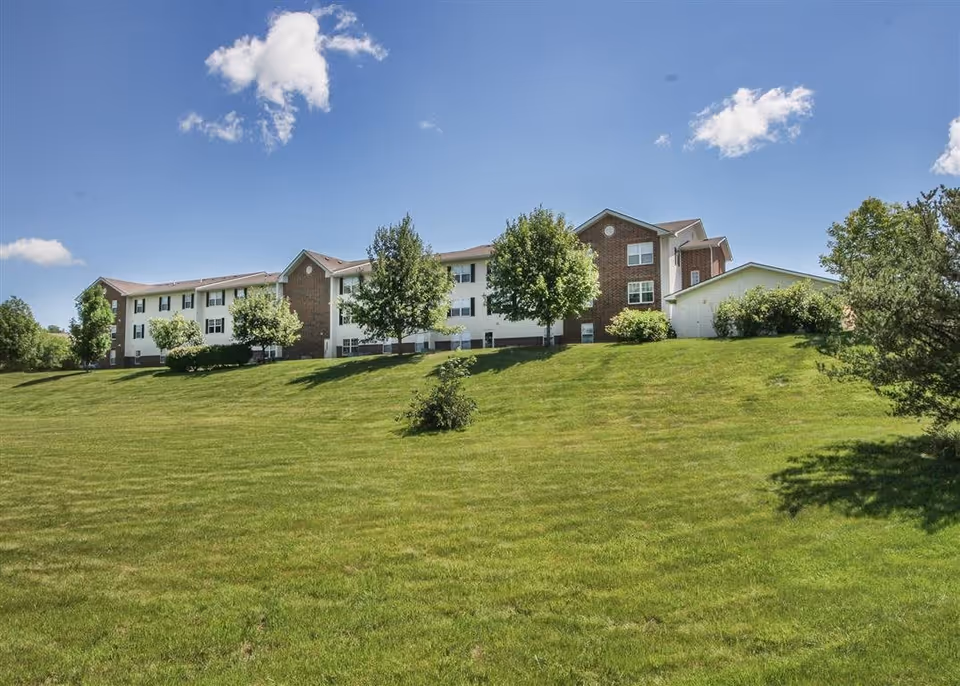 Exterior view of a multi-story senior living facility building with white and brick walls, surrounded by green grass and trees under a blue sky with a few clouds.