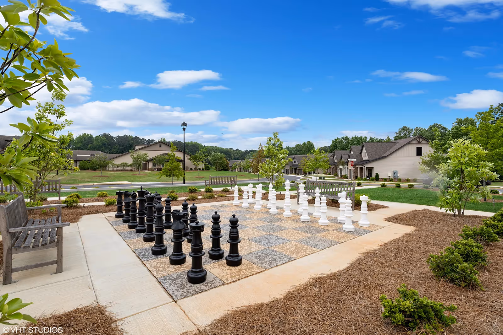 Outdoor area featuring a large chessboard with oversized black and white chess pieces set up for a game. Surrounding the chessboard are wooden benches, small trees, and landscaped greenery. In the background, there are residential buildings under a blue sky with scattered clouds.