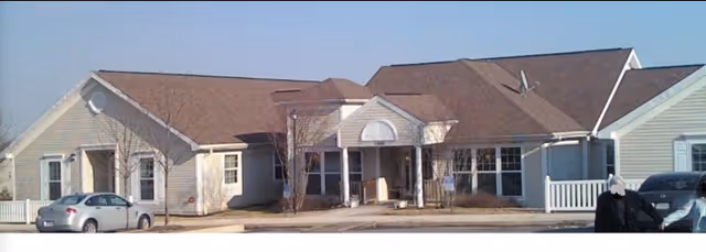 Front exterior view of a single-story senior living facility building with beige siding, multiple peaked roofs, a covered entrance with white columns, several windows, a few parked cars, and a clear blue sky.