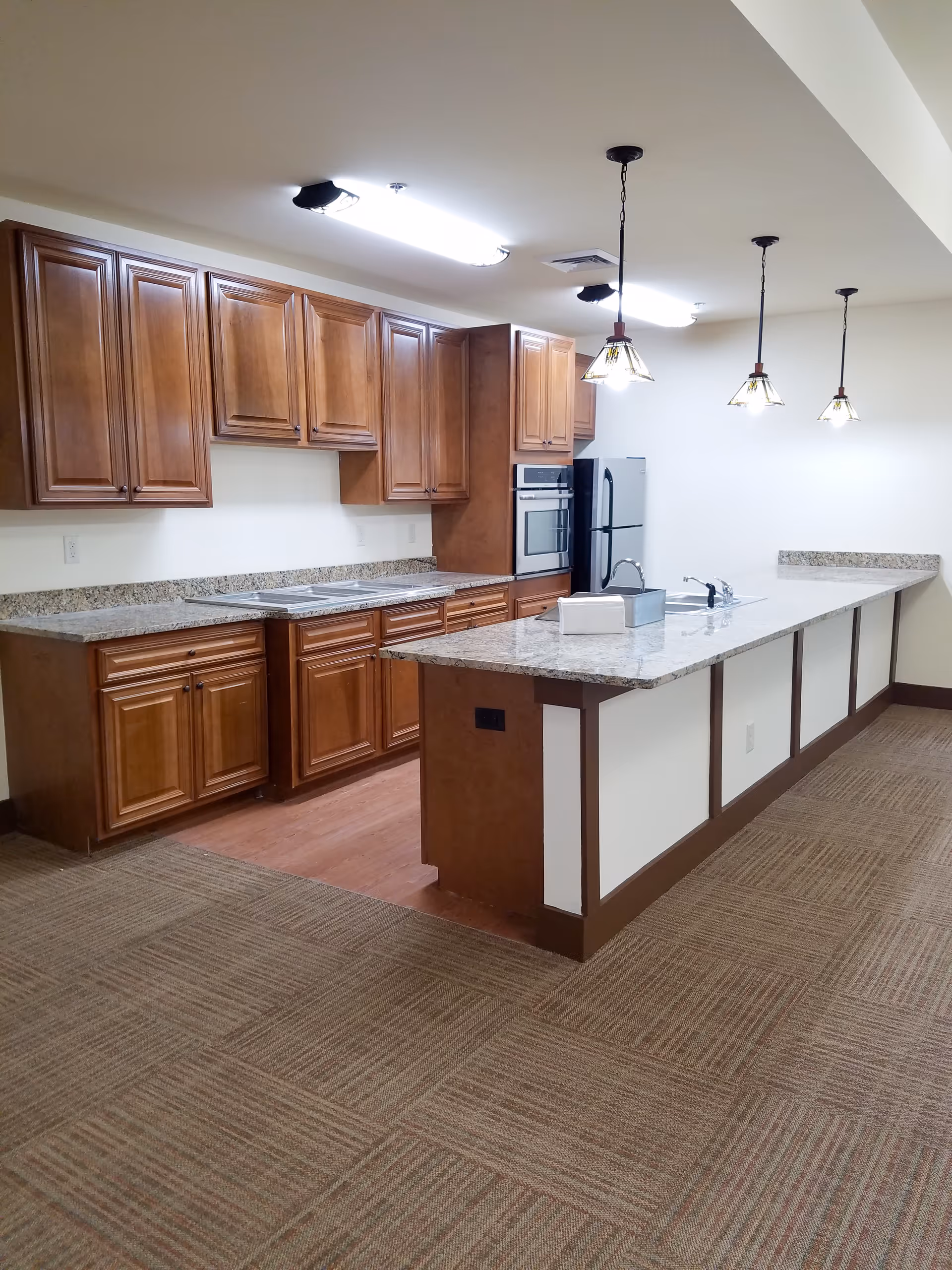 A spacious kitchen area with wooden cabinets, granite countertops, and a large island with a sink. The kitchen features a refrigerator, built-in oven, and three pendant lights hanging above the island. The floor is carpeted with a patterned design, and the walls are painted white.