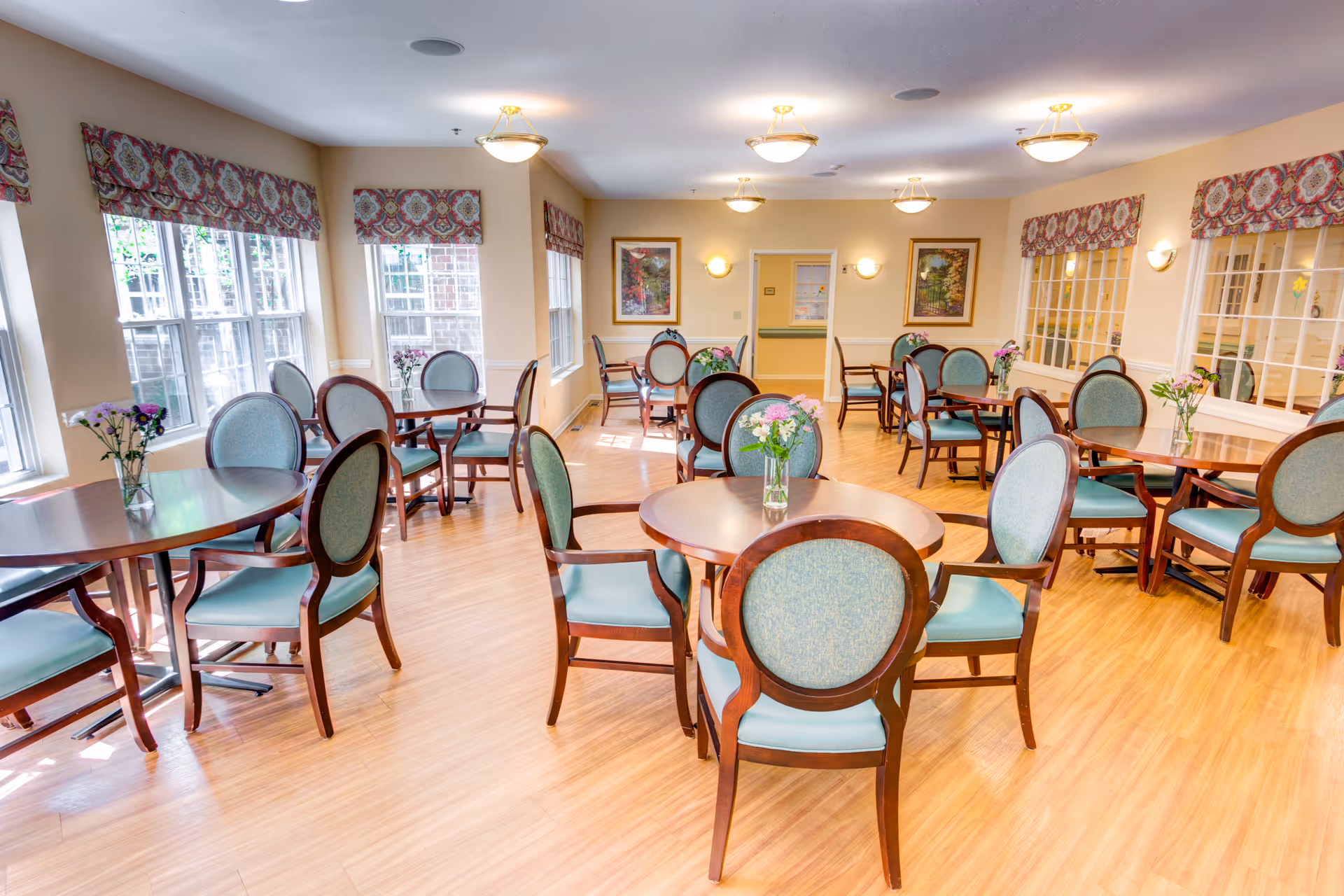 Bright dining room with multiple round wooden tables and cushioned chairs with blue upholstery. Each table has a small vase with flowers. Large windows with patterned valances allow natural light to fill the room. The walls are painted a soft beige, and framed artwork hangs on the far wall.