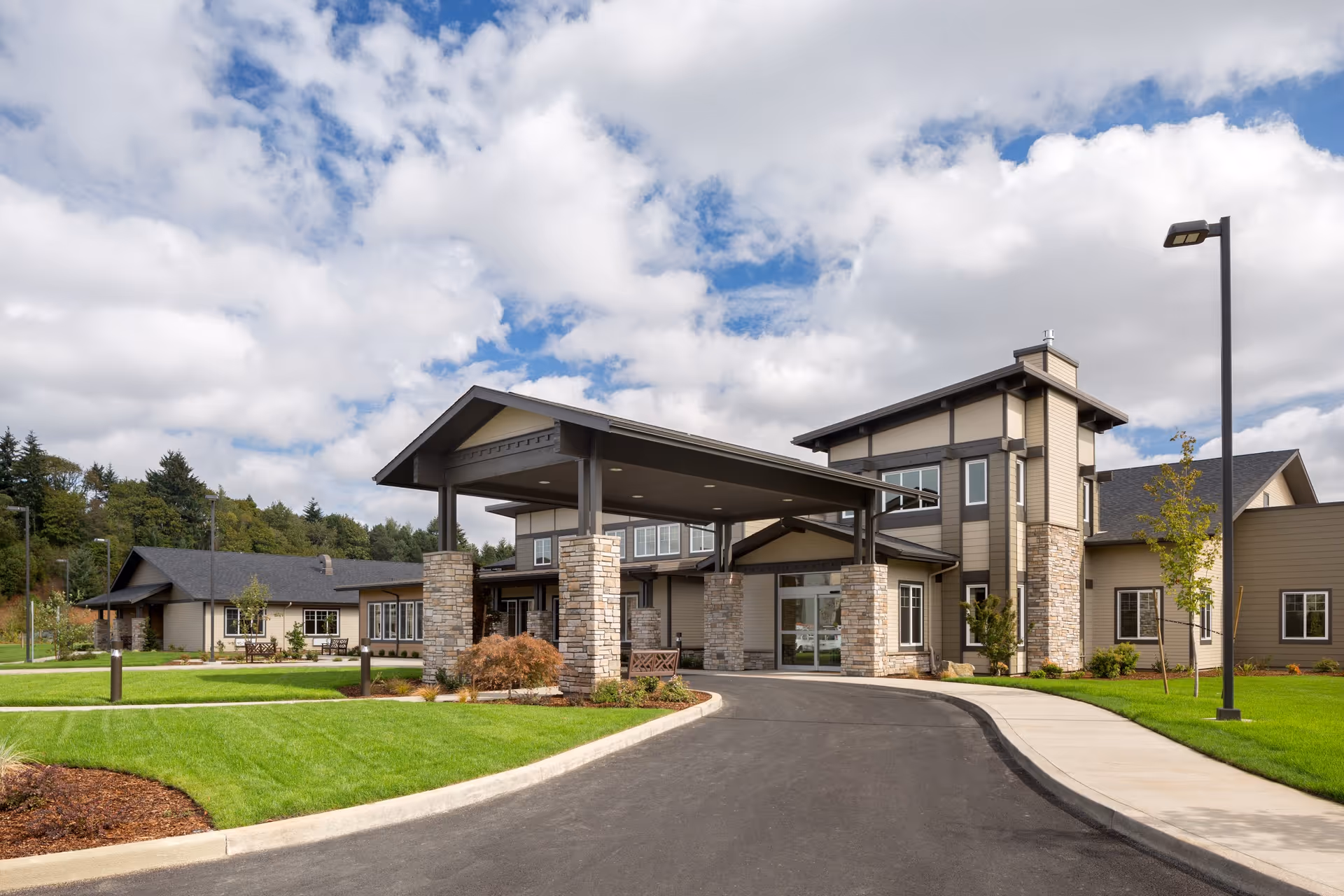 Front entrance of a senior living facility with a covered porte-cochere, stone columns, and landscaped lawns under a cloudy sky.