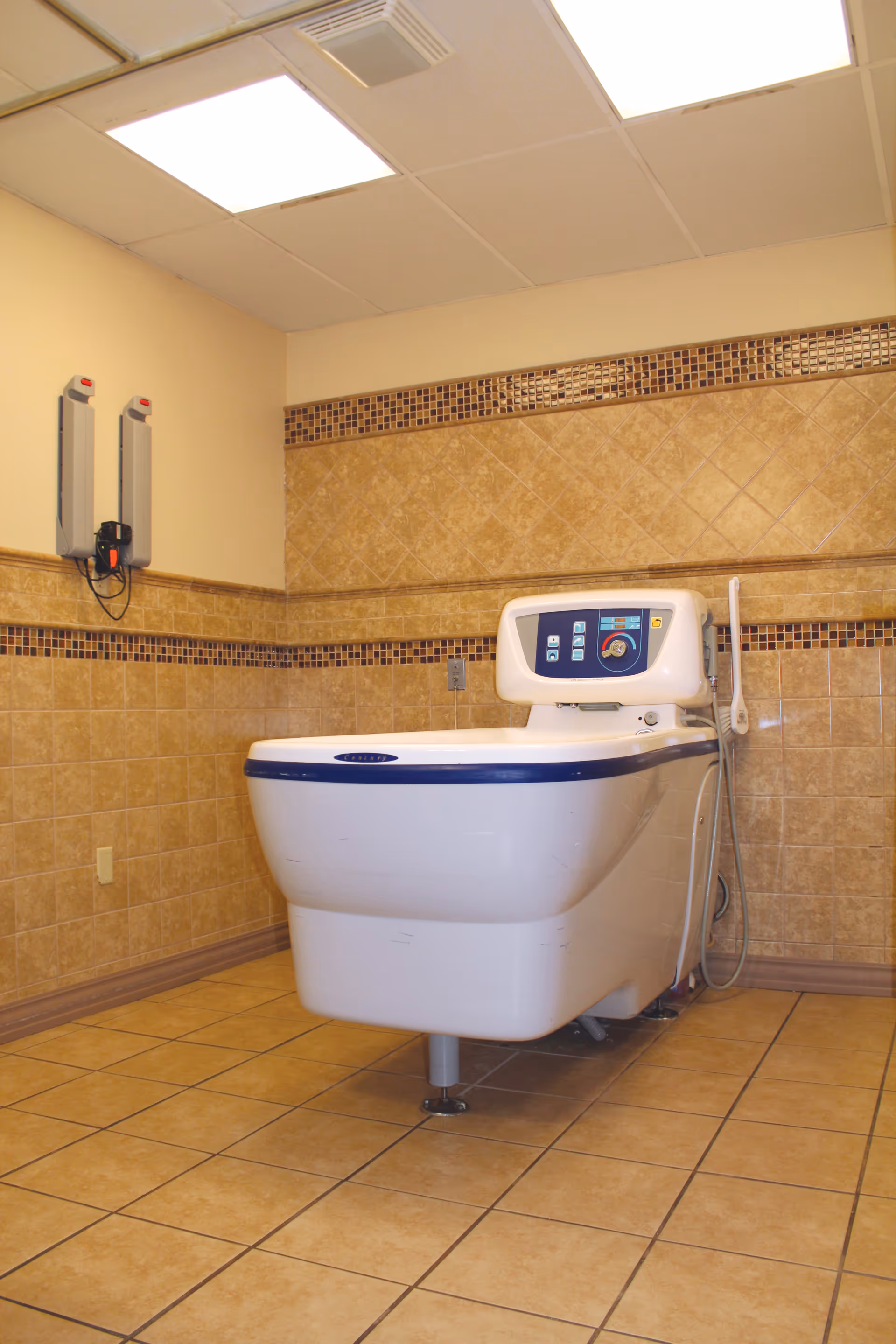 A medical bathing tub with control panel in a tiled room with beige walls and floor, designed for assisted bathing in a nursing center.