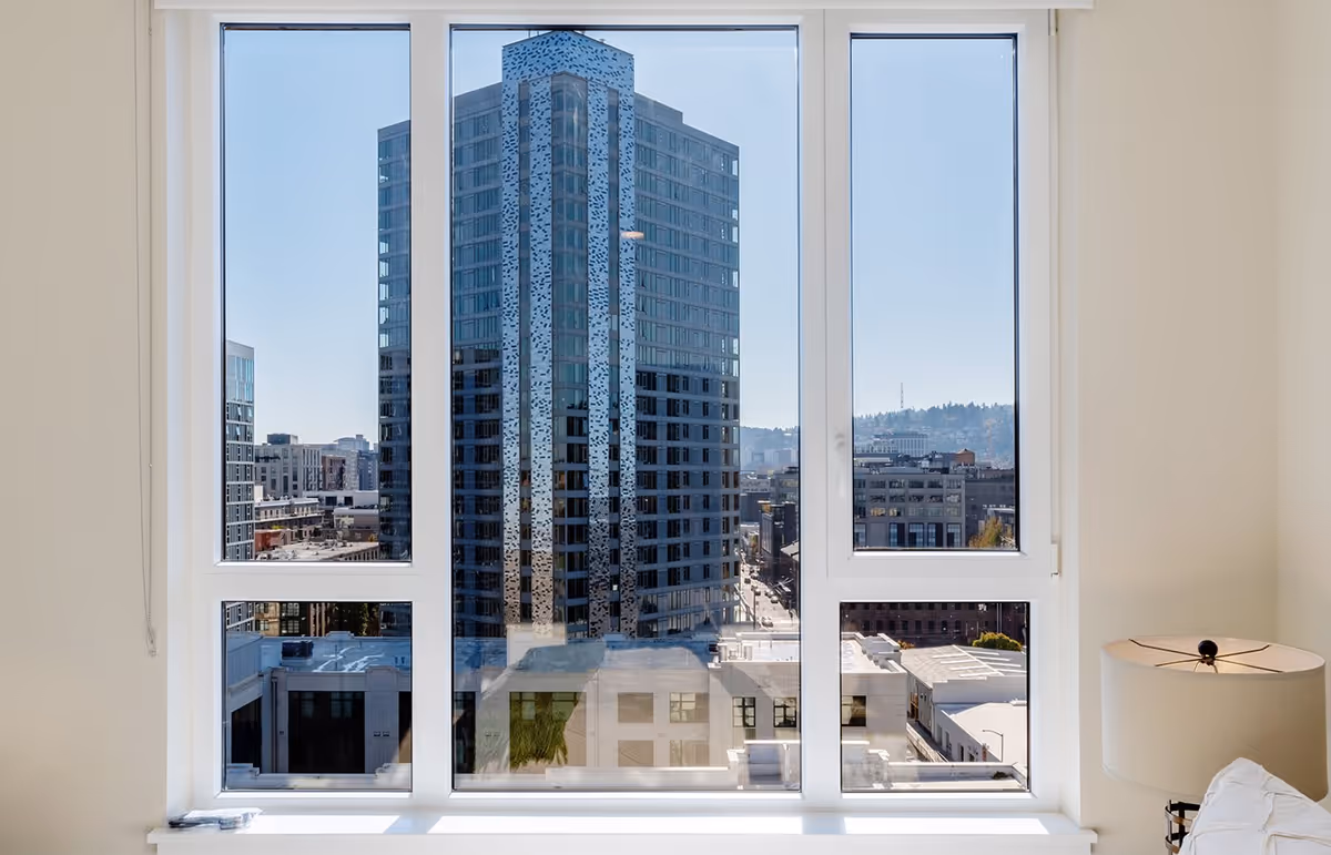 A tall glass office building and city skyline viewed through a large apartment window with a lamp on the right.
