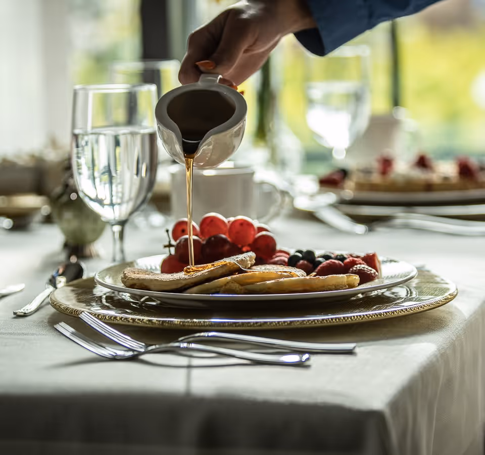 A close-up of a breakfast table setting with a plate of pancakes and fresh berries. A hand is pouring syrup from a small white pitcher onto the pancakes. The table is set with silverware, glasses of water, and a white tablecloth, with a softly blurred background showing more table settings and natural light coming through windows.