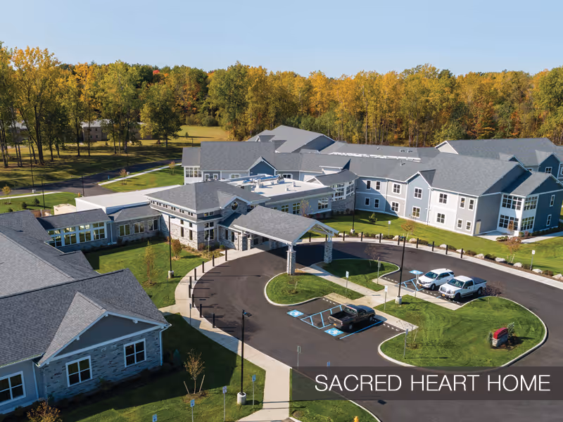 Aerial view of a large gray senior living building with a covered entrance, circular driveway, parked cars, and surrounding trees.