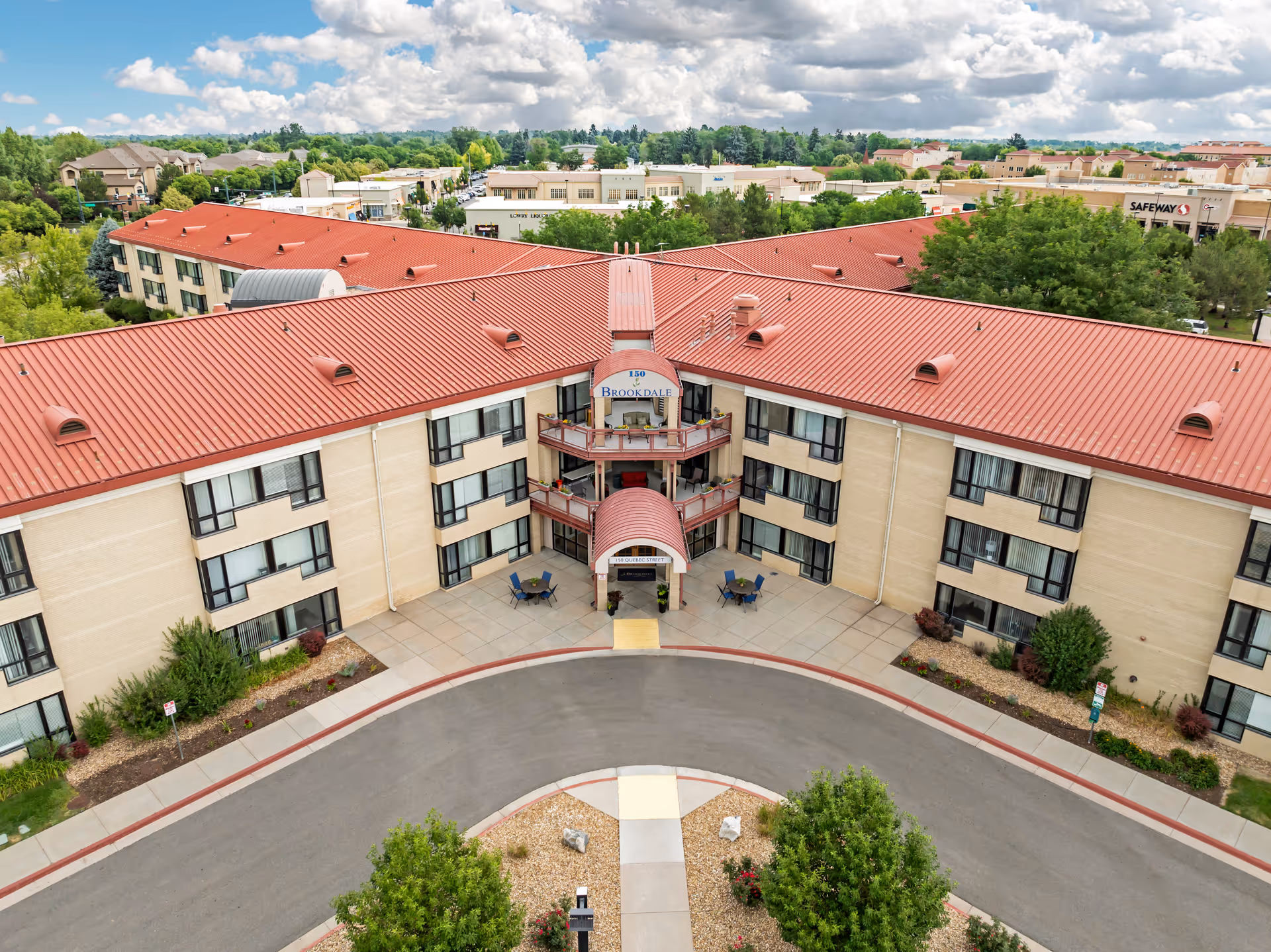 Aerial view of the Brookdale Lowry senior living facility showing a large building with a red roof and beige walls, surrounded by trees and greenery. The entrance features a covered walkway with the Brookdale sign and outdoor seating areas. The surrounding area includes other buildings and a Safeway store in the background under a partly cloudy sky.