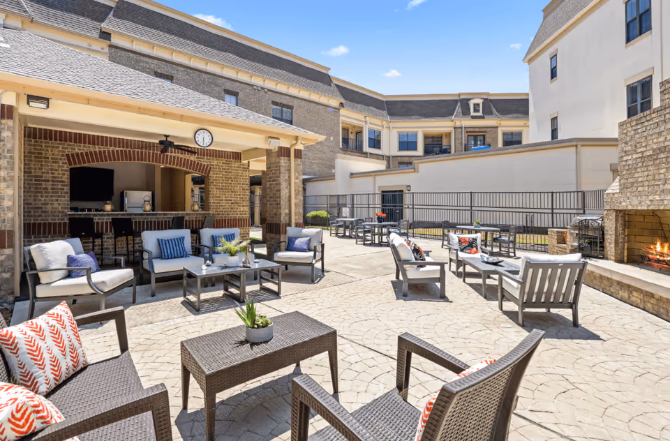 Outdoor patio area at Discovery Village At Castle Hills featuring multiple seating arrangements with cushioned chairs and tables, a covered bar area with stools, a wall-mounted clock, and a fireplace with a fire burning. The space is surrounded by a multi-story building under a clear blue sky.