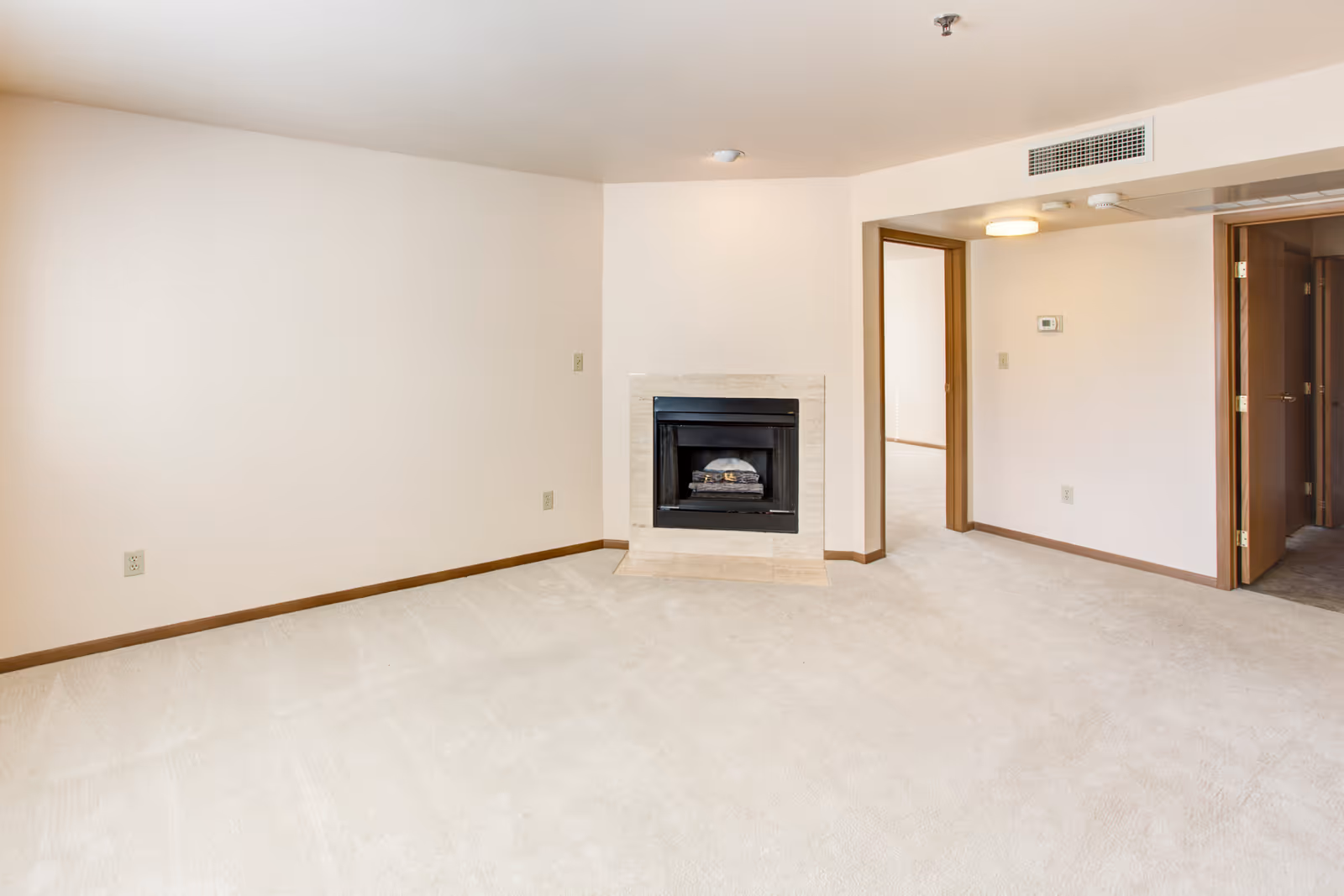 Empty carpeted living room featuring a central gas fireplace, beige walls, and doorways to adjoining rooms.