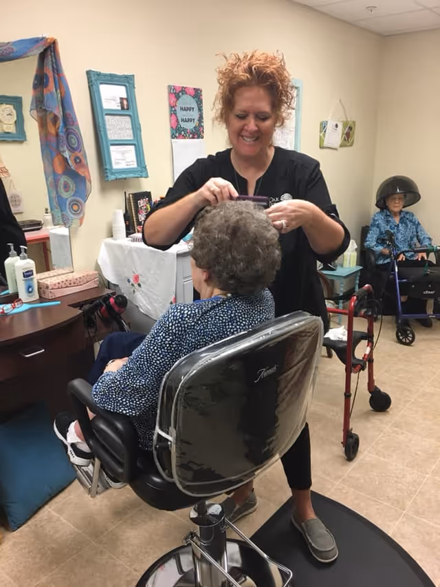 A woman with curly red hair is styling the hair of an elderly woman seated in a salon chair inside a room. Another elderly woman is sitting under a hair dryer in the background. The room has a table with various items, framed pictures on the wall, and a walker nearby.