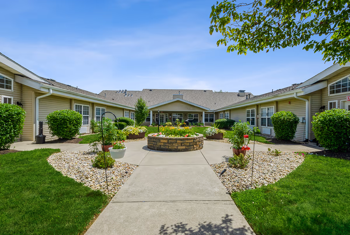 Outdoor courtyard area of Worthington Place senior living facility with a circular flower bed in the center, surrounded by a concrete walkway, green bushes, and beige buildings under a clear blue sky.