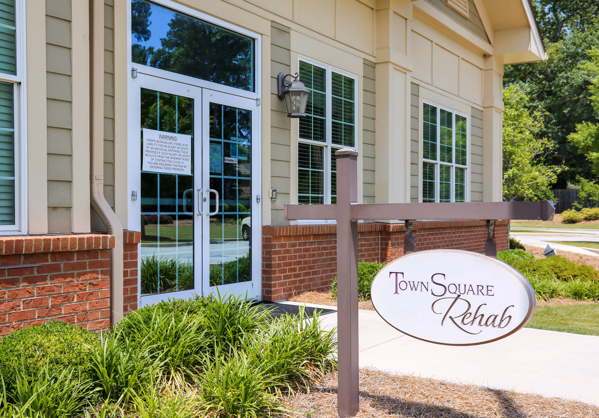 Front entrance of a rehab facility with glass double doors, large windows, landscaping, and a hanging 'Town Square Rehab' sign.