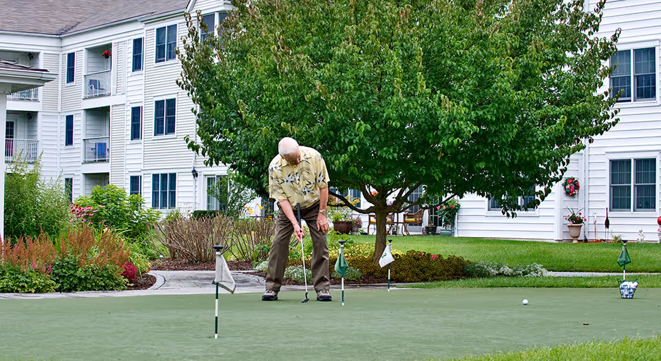 An elderly man playing golf on a putting green surrounded by landscaped gardens and a large tree, with a white multi-story residential building in the background.