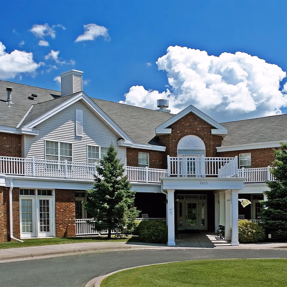 Exterior front view of a two-story senior living facility building with brick and white siding, a covered entrance with white columns, balconies with white railings, and a well-maintained lawn and trees under a blue sky with scattered clouds.