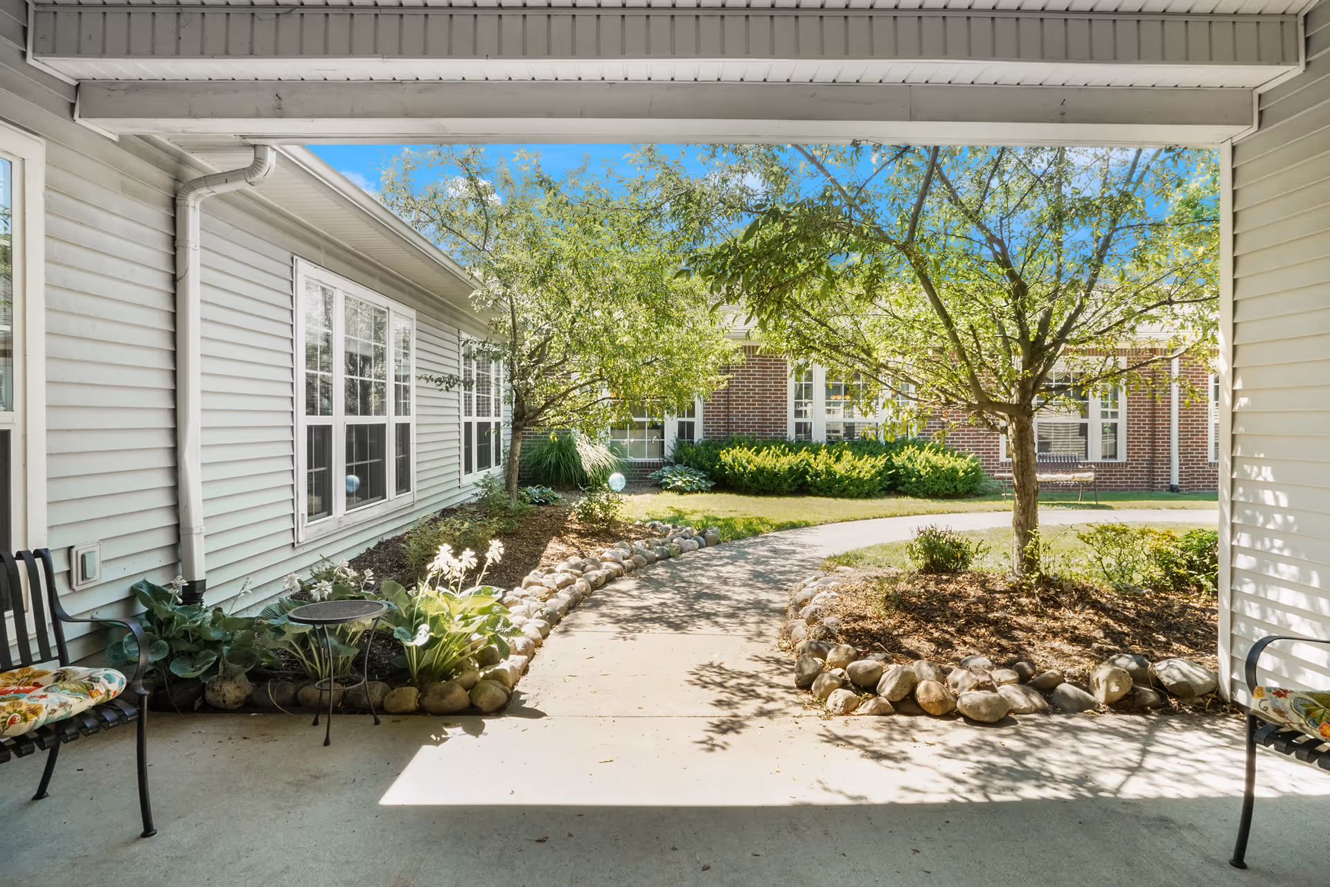 View from a covered patio area looking out onto a landscaped garden path with trees, shrubs, and a brick building in the background. There are two chairs with floral cushions on either side of the patio.