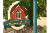 A decorative outdoor sign for Home 2 Me Inc featuring a red house with a large window, surrounded by greenery and mounted on a green post.