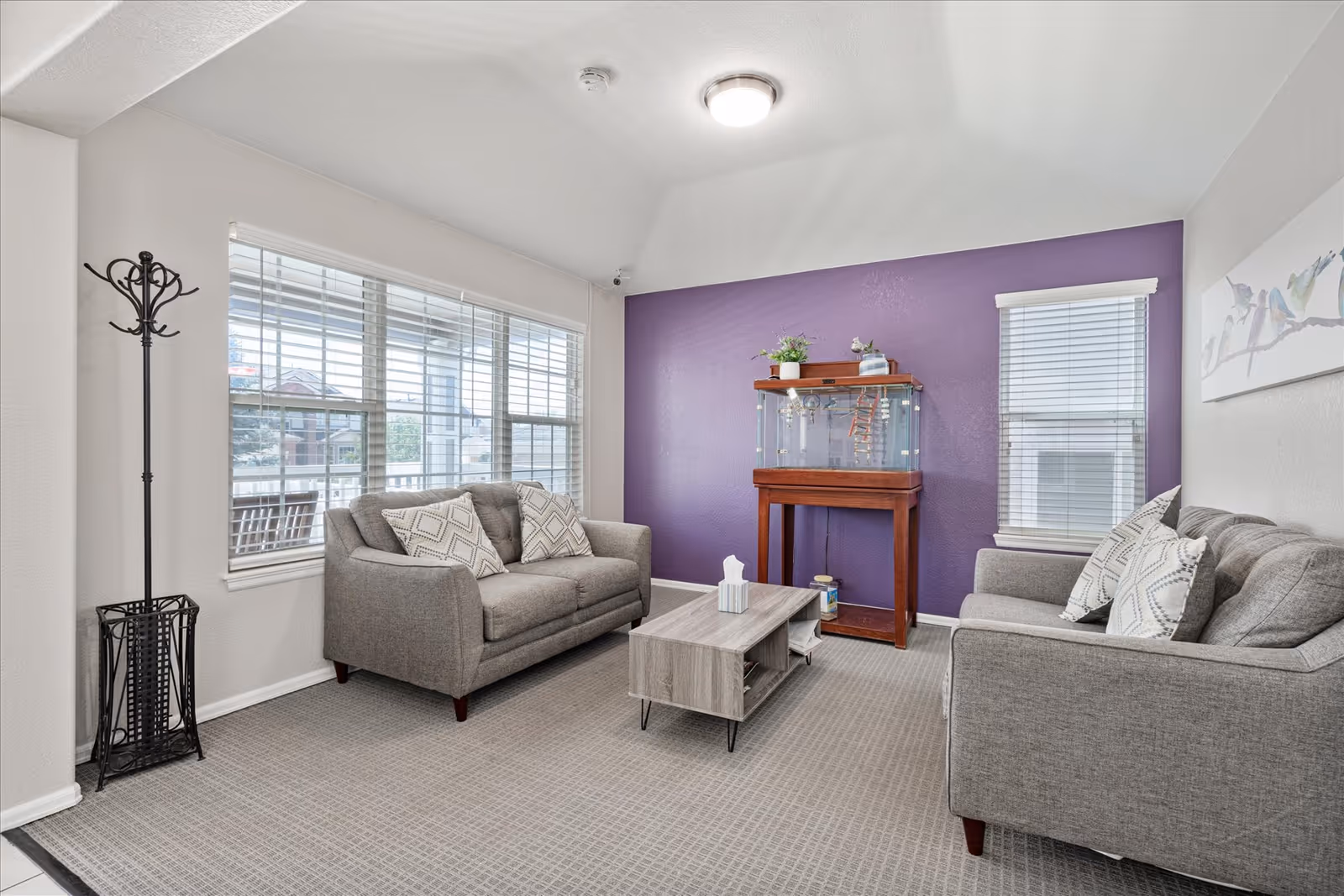 A bright living room with two gray sofas facing each other, each with patterned throw pillows. A small wooden coffee table is placed between the sofas. The room has a large window with white blinds on one side and a smaller window with blinds on the adjacent wall. A purple accent wall features a wooden fish tank stand with a fish tank on top and some decorative items. A coat rack stands near the large window, and a piece of artwork hangs on the white wall above one sofa.