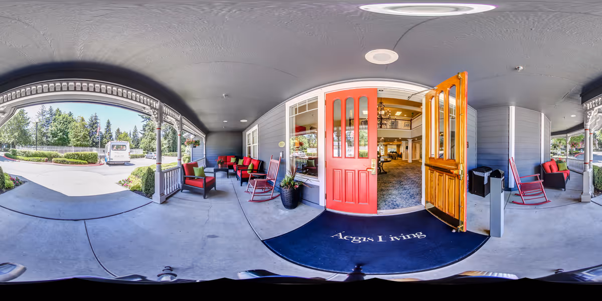Wide view of the entrance to Aegis Living Kirkland showing an outdoor covered porch area with red cushioned chairs and rocking chairs. The porch leads to double doors, one red and one wooden, opening into a spacious interior with carpeted floors and seating areas. Outside, there are trees, bushes, and a driveway with a parked vehicle.