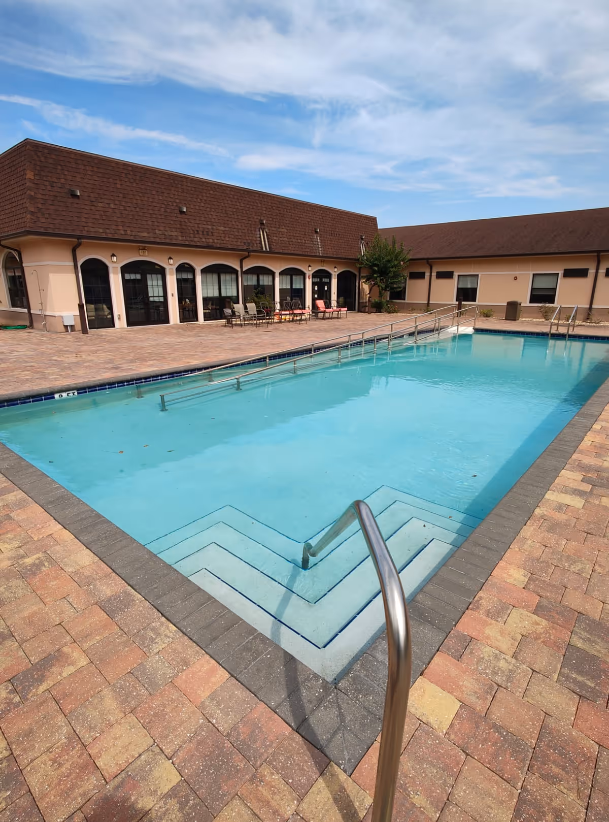 A rectangular outdoor swimming pool with steps and metal handrails surrounded by brick pavers and patio seating in front of a single-story building.
