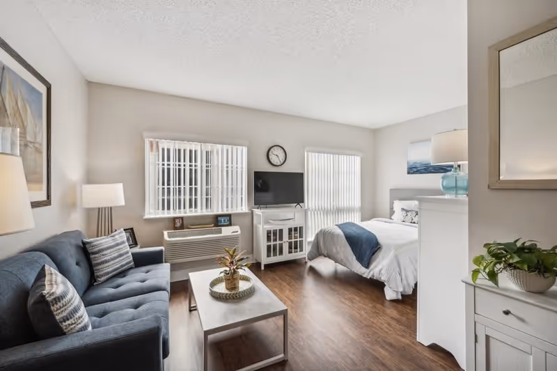 A bright and cozy studio apartment with a blue sofa and patterned pillows on the left, a coffee table with a plant in the center, and a bed with white and blue bedding on the right. There is a TV on a white stand against the wall between two windows with vertical blinds. A clock hangs above the TV. The floor is wooden, and there are lamps and framed artwork decorating the room.