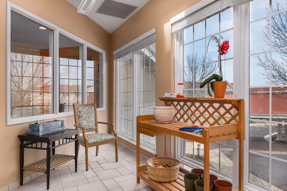 Bright sunroom with large windows, a wooden potting bench holding an orchid and plant pots, a patterned armchair and small side table.