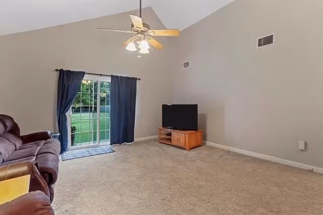 A spacious living room with beige carpet and light gray walls. There is a ceiling fan with lights hanging from a vaulted ceiling. A sliding glass door with dark blue curtains leads outside to a green lawn. A brown leather recliner is partially visible on the left, and a flat-screen TV sits on a wooden stand against the right wall.