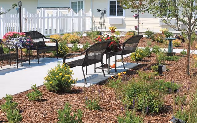 Sunlit outdoor courtyard with metal benches, flower planters, a paved walkway, and a white picket fence in front of a light-colored building.