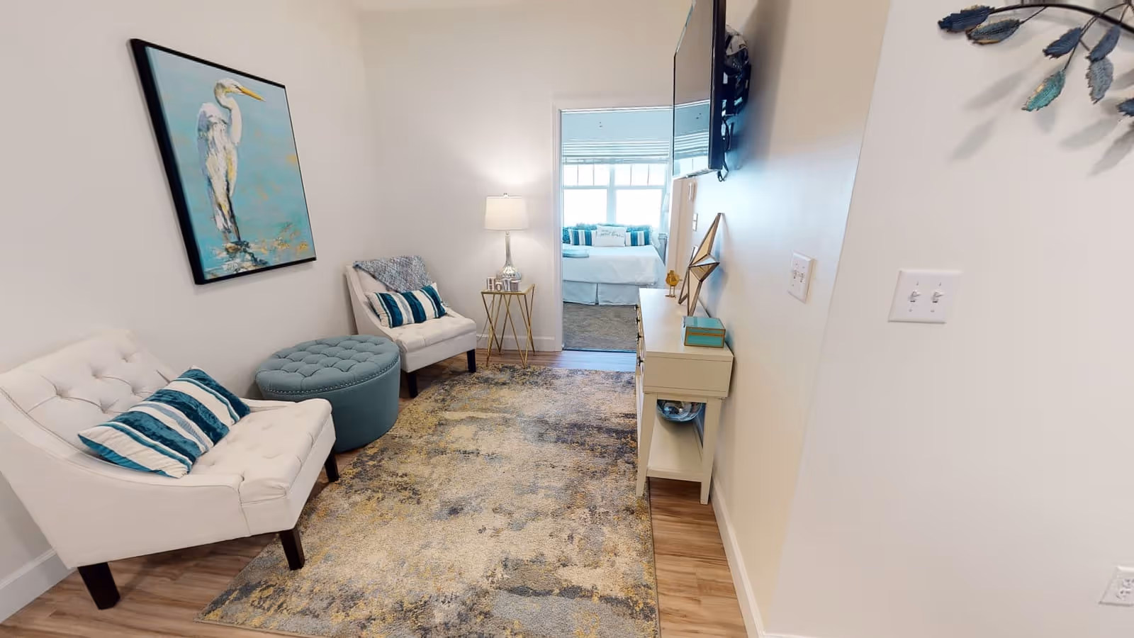 A small living room area with two white cushioned chairs, a teal ottoman, and a patterned rug on a wooden floor. A painting of a white bird on a blue background hangs on the left wall. A small side table with a lamp and decorative items is placed between the chairs. A TV is mounted on the right wall above a console table with decorative items. Through an open doorway, a bedroom with a bed and pillows is visible.