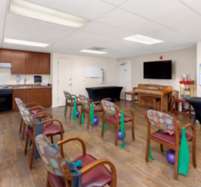 A spacious room with wooden flooring and several chairs arranged in rows. Each chair has exercise equipment including green resistance bands and purple exercise balls placed nearby. The room has a kitchenette area with cabinets, a stove, and a sink on one side. There is a piano against the far wall beneath a mounted flat-screen TV, and a table covered with a black cloth is also visible. The ceiling has recessed lighting and a whiteboard is mounted on the wall near the doors.