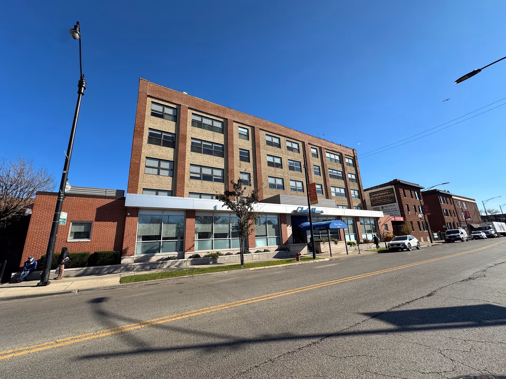 Exterior street view of Clark Manor, a multi-story brick building with a front entrance awning and parked cars.